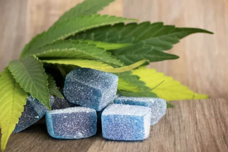 Blue sugar-coated cannabis gummy edibles beside green cannabis leaves on a wooden table